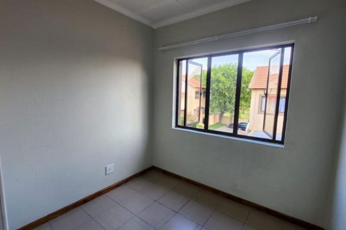A clean, empty bedroom featuring tiled flooring, neutral walls, and a large casement window providing natural light and a view of the complex.