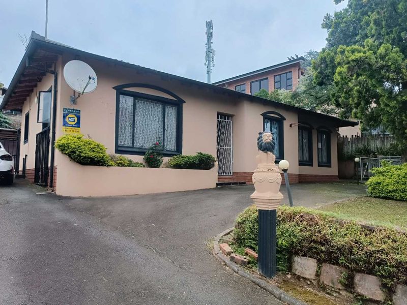 Front elevation of a single-story residential house featuring an asphalt driveway, security gate, and established garden area with a decorative pillar.