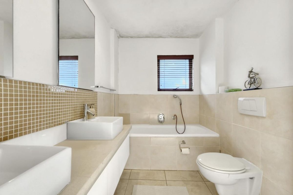 A functional bathroom featuring a double vanity, wall-mounted toilet, and a built-in bathtub with neutral tiling and a mosaic backsplash.