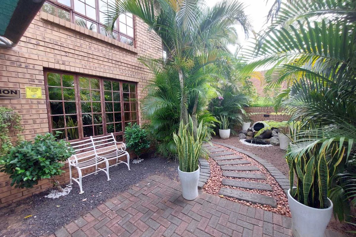 A well-maintained rear garden area featuring a brick facade, paved pathways, established palm trees, and a decorative stone water feature.