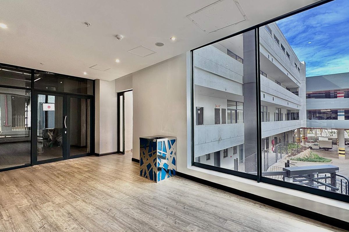 Modern office space featuring large floor-to-ceiling windows, wood-look flooring, and recessed ceiling lighting with views of the surrounding office complex.