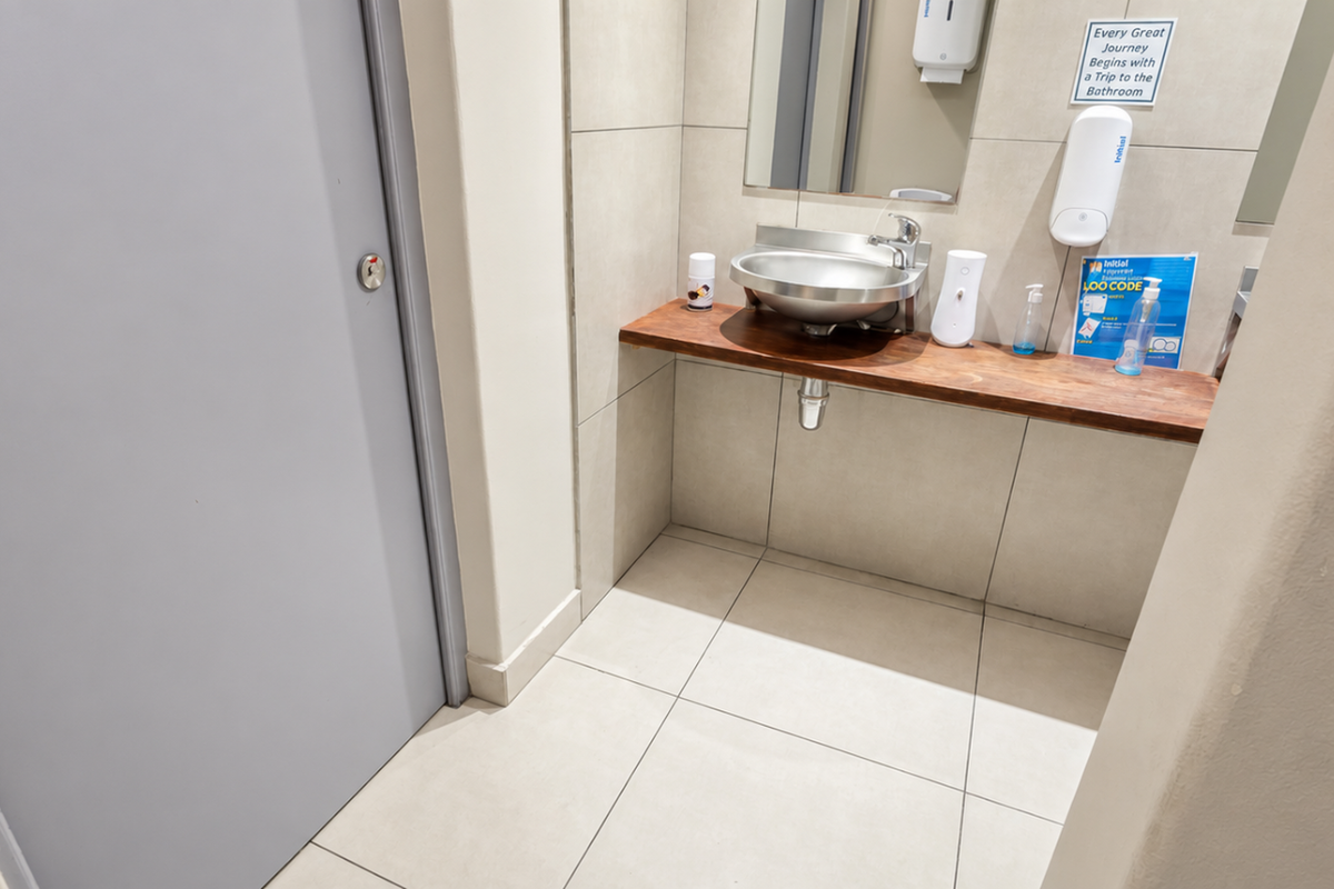 A clean, modern office bathroom featuring a stainless steel basin set into a wooden vanity countertop with neutral tiled walls and flooring.