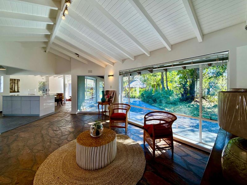 Spacious open-plan living area featuring vaulted ceilings with exposed beams, flagstone flooring, and large windows providing abundant natural light and garden views.