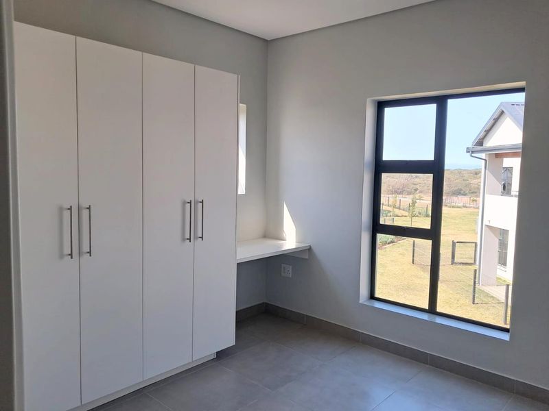 A bright, modern bedroom featuring built-in white cupboards, a fitted desk area, tiled flooring, and large windows with a pleasant exterior view.
