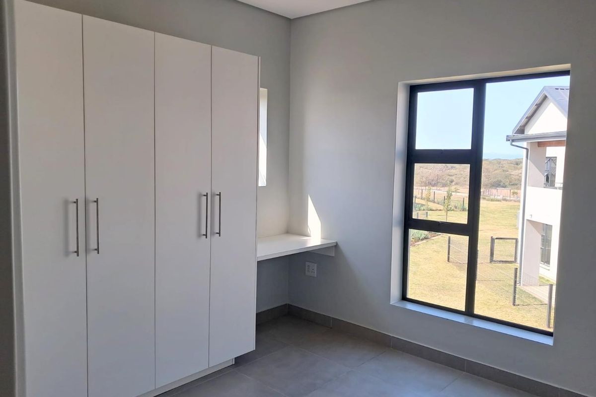 A bright, modern bedroom featuring built-in white cupboards, a fitted desk area, tiled flooring, and large windows with a pleasant exterior view.