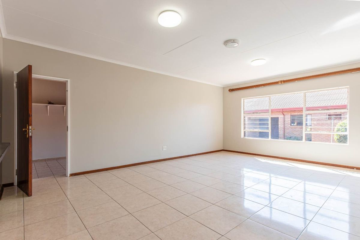 A spacious, empty living area featuring neutral tiled flooring, large windows, and direct access to an adjacent room.