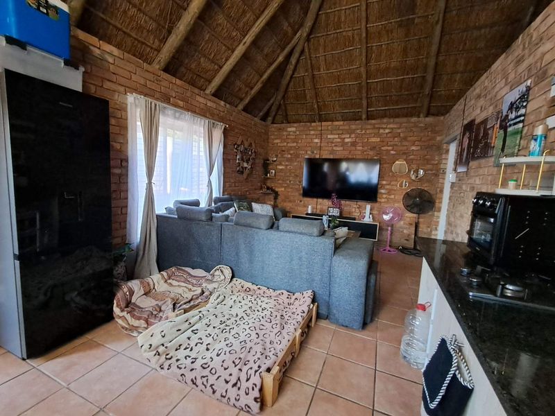 Open-plan living area featuring exposed brick walls, a traditional thatched roof, and tiled flooring, combining kitchen and lounge spaces in a rustic cottage style.