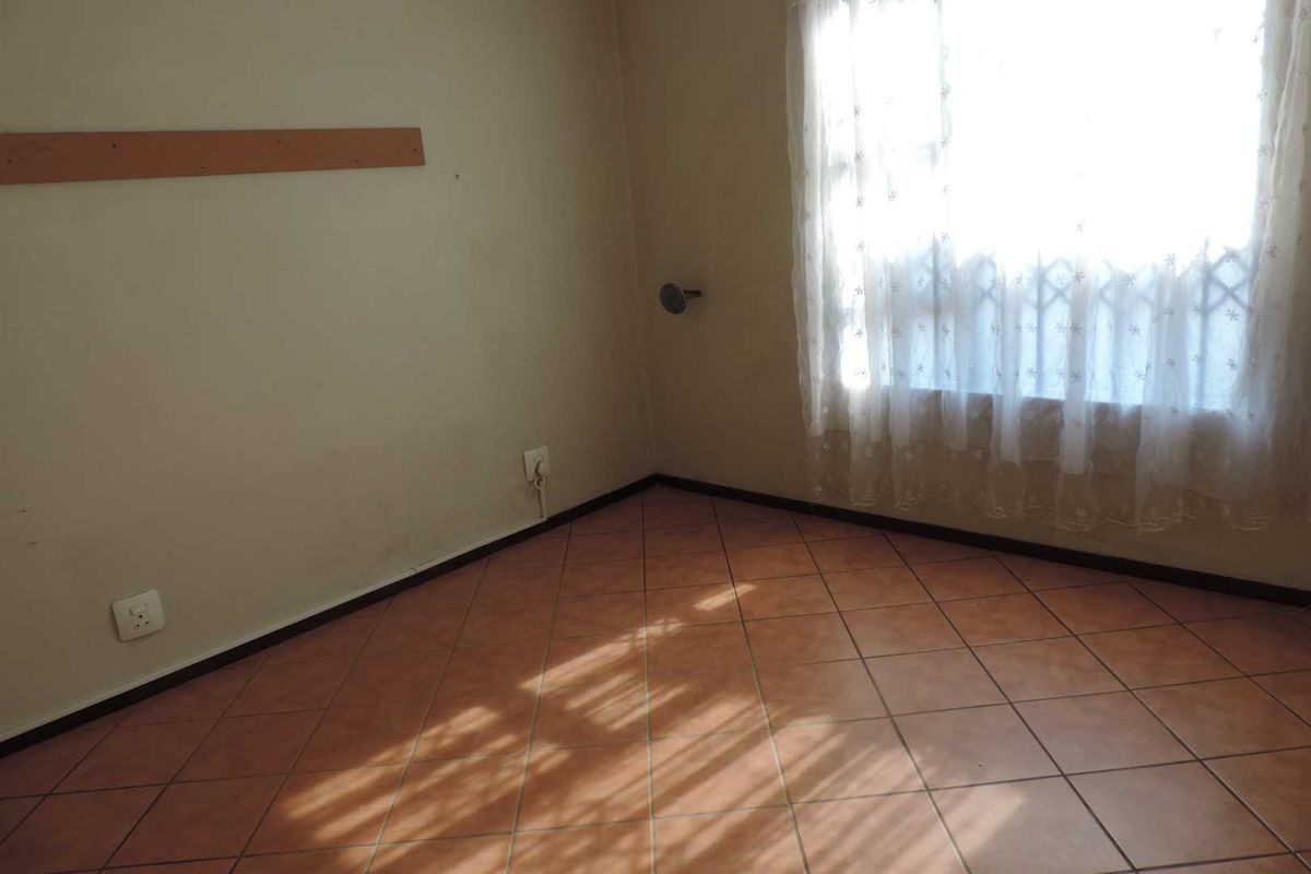 A vacant bedroom featuring terracotta floor tiles, a window with security bars, and neutral wall finishes.