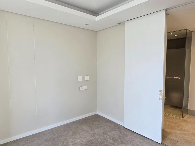 A clean, modern bedroom featuring neutral walls, plush carpeting, a recessed ceiling, and a sliding door leading to an adjacent bathroom.