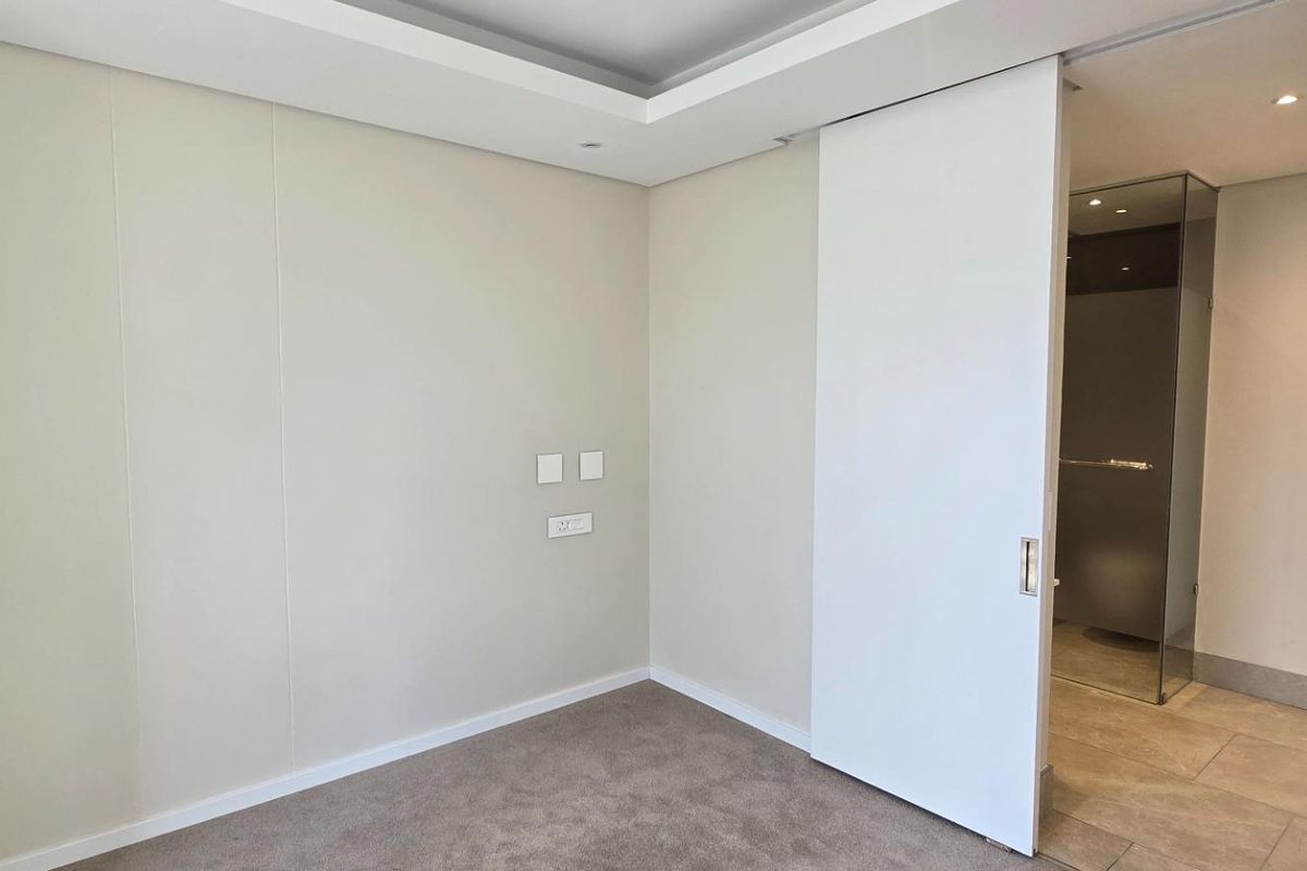 A clean, modern bedroom featuring neutral walls, plush carpeting, a recessed ceiling, and a sliding door leading to an adjacent bathroom.