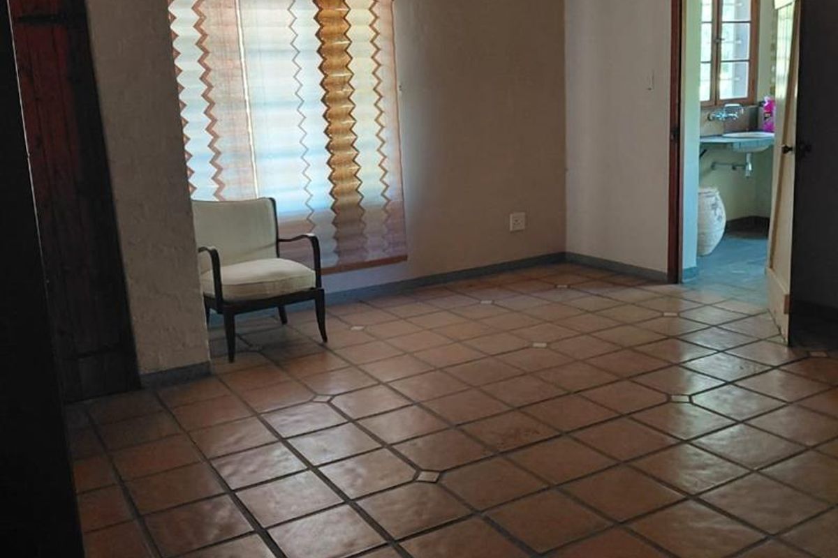 A spacious living area featuring traditional terracotta floor tiles and neutral wall finishes, with an adjacent room visible through the doorway.