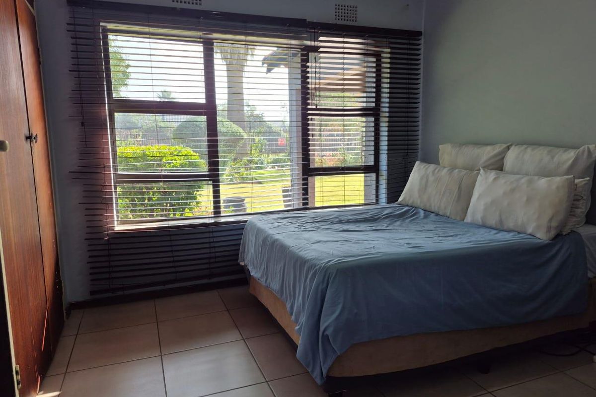 A bedroom featuring large windows with venetian blinds, tiled flooring, and built-in wooden cabinetry, offering a view of the garden.