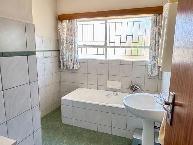 A functional bathroom featuring a pedestal washbasin, a built-in bathtub, and tiled finishes throughout, offering a classic layout with security window bars.