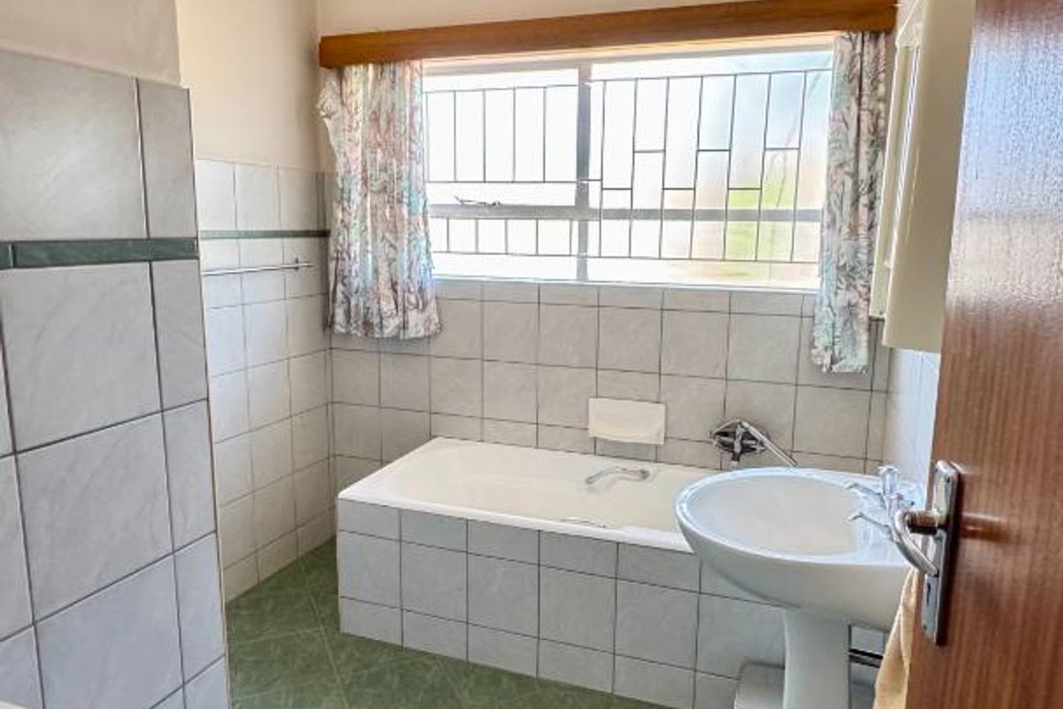 A functional bathroom featuring a pedestal washbasin, a built-in bathtub, and tiled finishes throughout, offering a classic layout with security window bars.