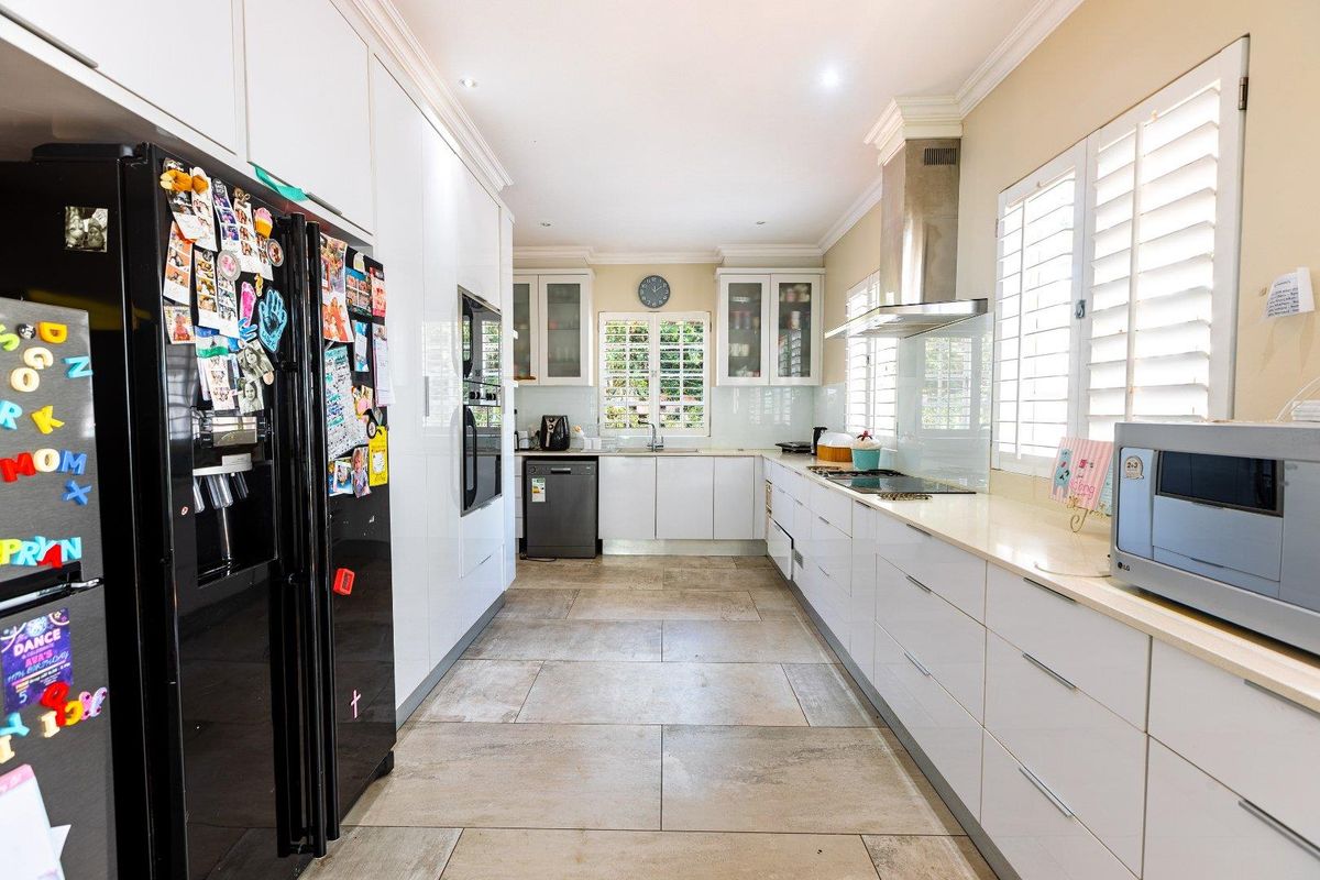 Modern galley-style kitchen featuring white cabinetry, stone countertops, integrated oven, stainless steel extractor fan, and tiled flooring with plantation shutters.
