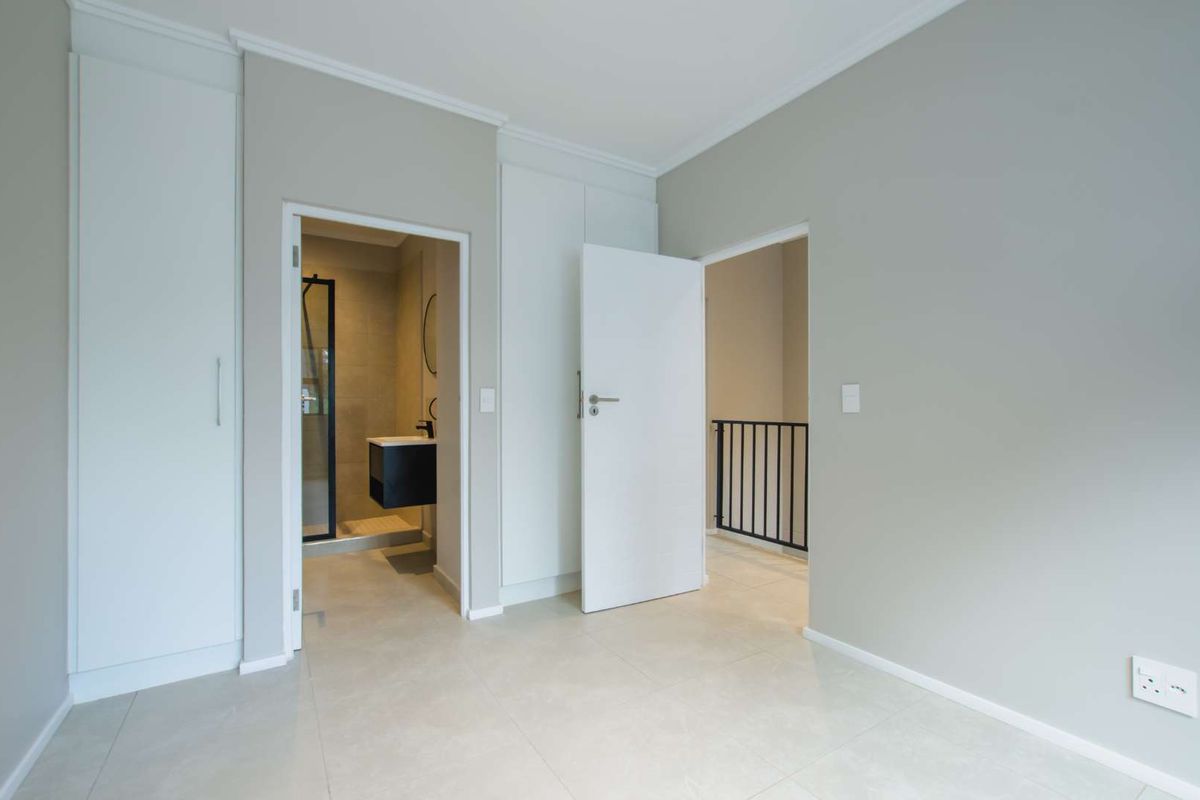 A bright, modern bedroom featuring neutral tiled flooring, built-in cabinetry, and direct access to an en-suite bathroom and hallway.