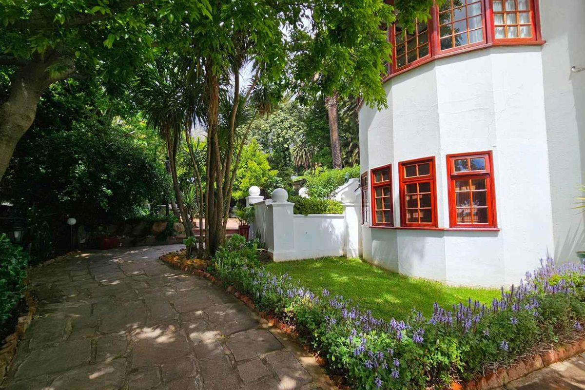 A well-maintained rear garden area featuring a stone paved pathway, lush greenery, and a white building facade with distinctive red-framed multi-pane windows.