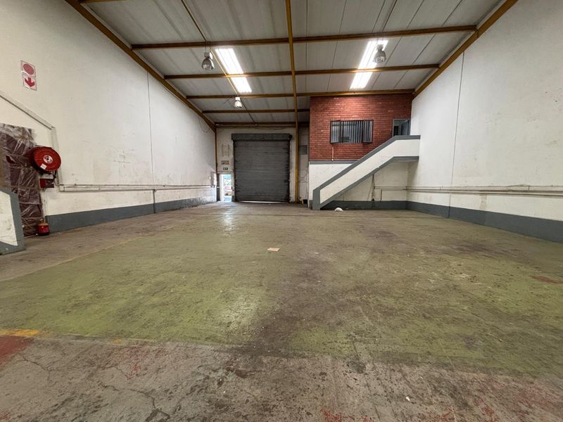 Spacious industrial warehouse unit featuring high ceilings, exposed steel trusses, a large roller shutter door, and a concrete floor with an internal staircase.