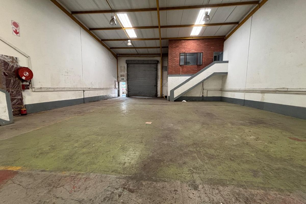 Spacious industrial warehouse unit featuring high ceilings, exposed steel trusses, a large roller shutter door, and a concrete floor with an internal staircase.