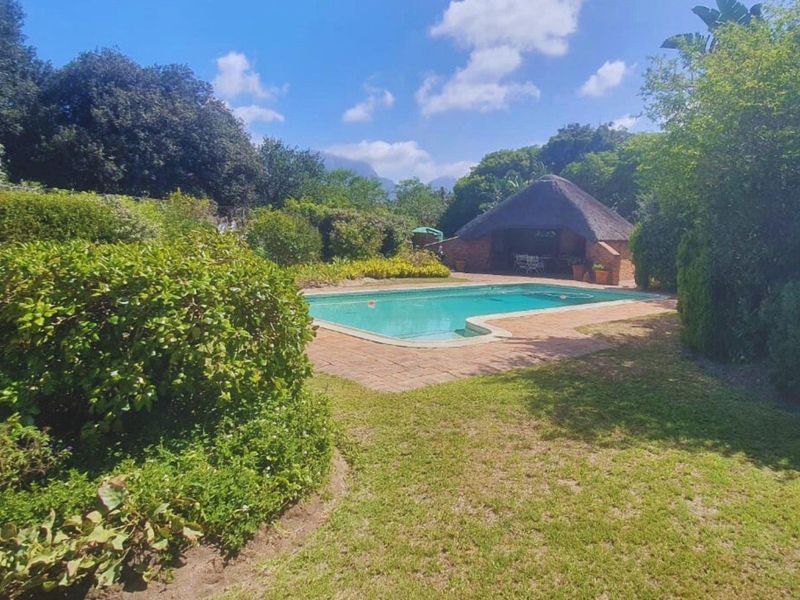 A private outdoor pool area featuring a swimming pool, brick-paved surround, and a traditional thatched lapa set within a lush, landscaped garden.