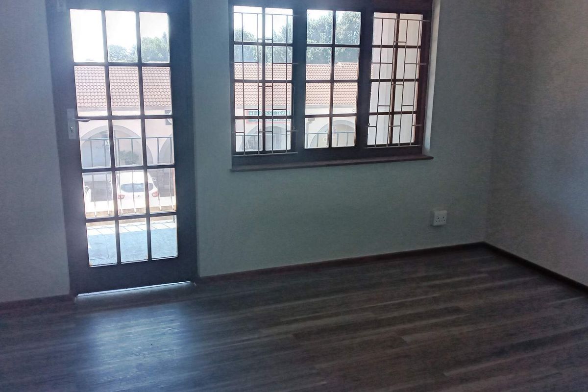 A bright, empty office space featuring laminate flooring, a glass-paneled french door, and windows fitted with security bars.