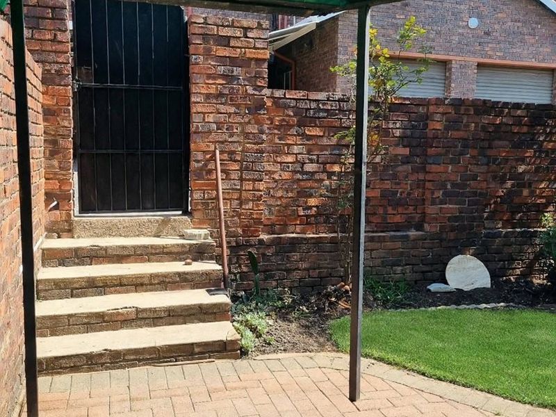 Outdoor patio area featuring brick paving, concrete steps, and a corrugated plastic roof structure, enclosed by a brick boundary wall.