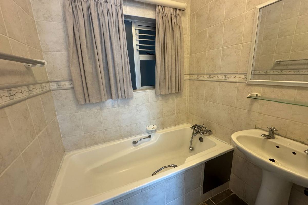 A functional bathroom featuring a built-in bathtub with shower mixer, pedestal basin, and wall-mounted mirror, finished with neutral ceramic wall tiles.