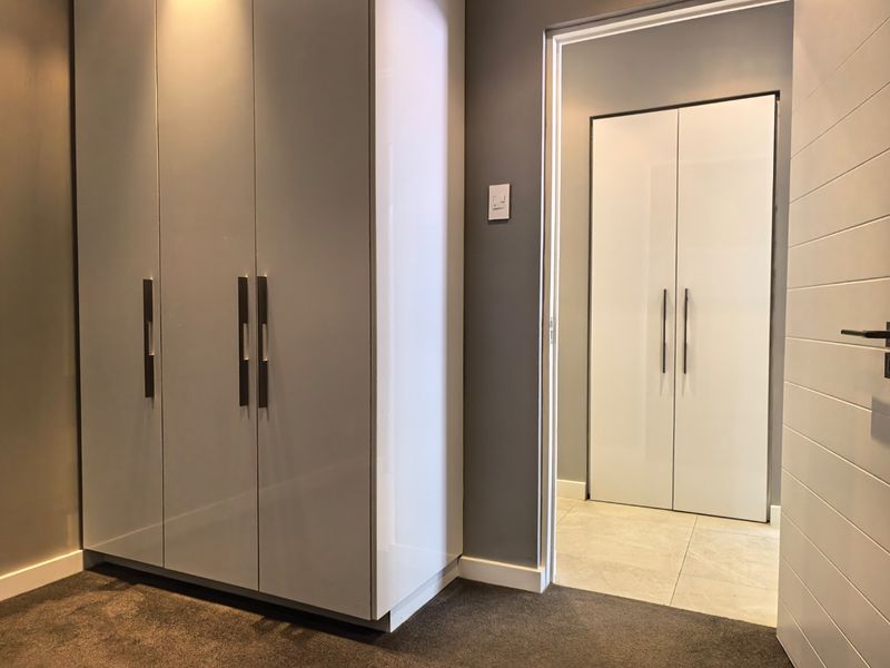 A modern hallway featuring high-gloss built-in cupboards, neutral grey walls, and a combination of tiled and carpeted flooring leading to an adjacent room.