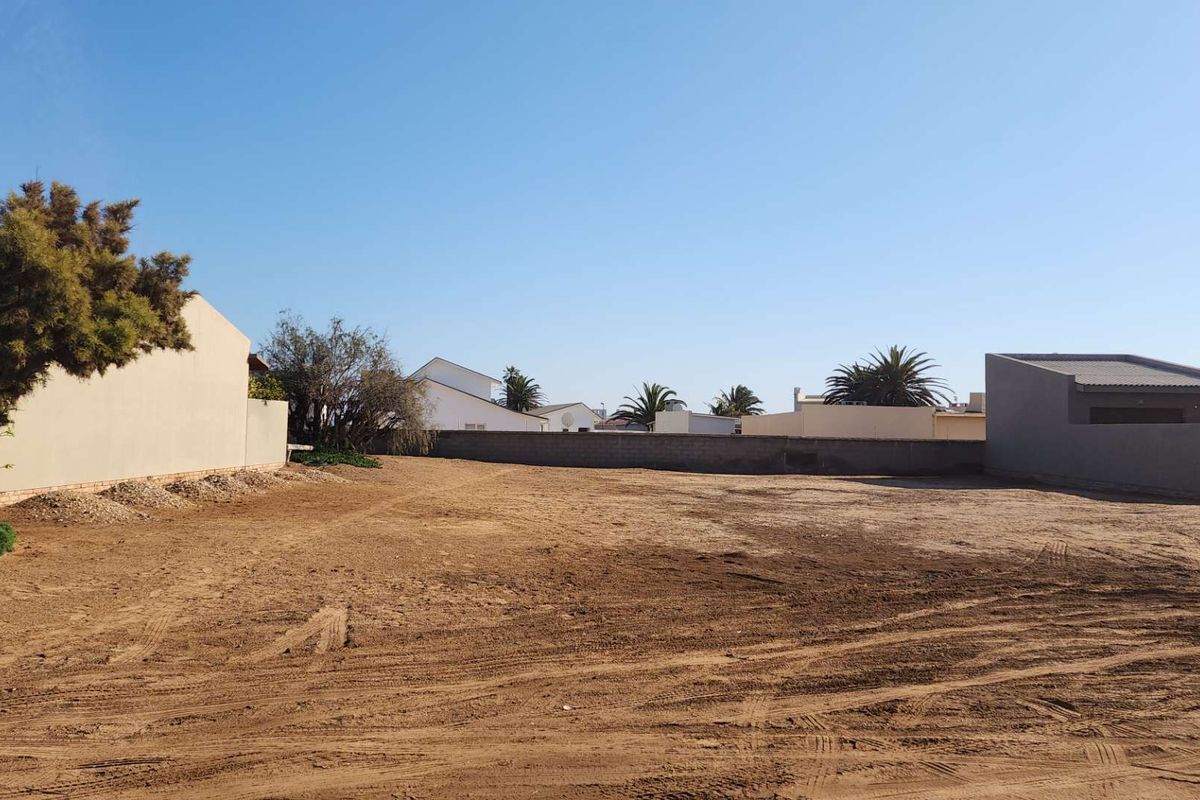 A vacant residential plot of land featuring cleared sandy ground, enclosed by boundary walls, and situated within an established residential neighborhood.