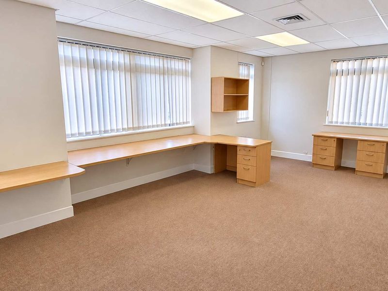 A spacious, well-lit office area featuring built-in wooden desks, integrated shelving, neutral carpeting, and large windows with vertical blinds.