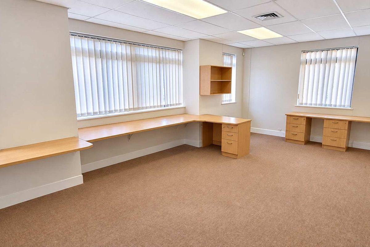 A spacious, well-lit office area featuring built-in wooden desks, integrated shelving, neutral carpeting, and large windows with vertical blinds.