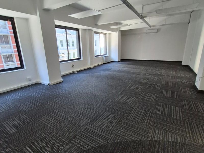 A spacious, open-plan office area featuring modern carpet tiles, exposed ceiling beams, and large windows providing ample natural light.