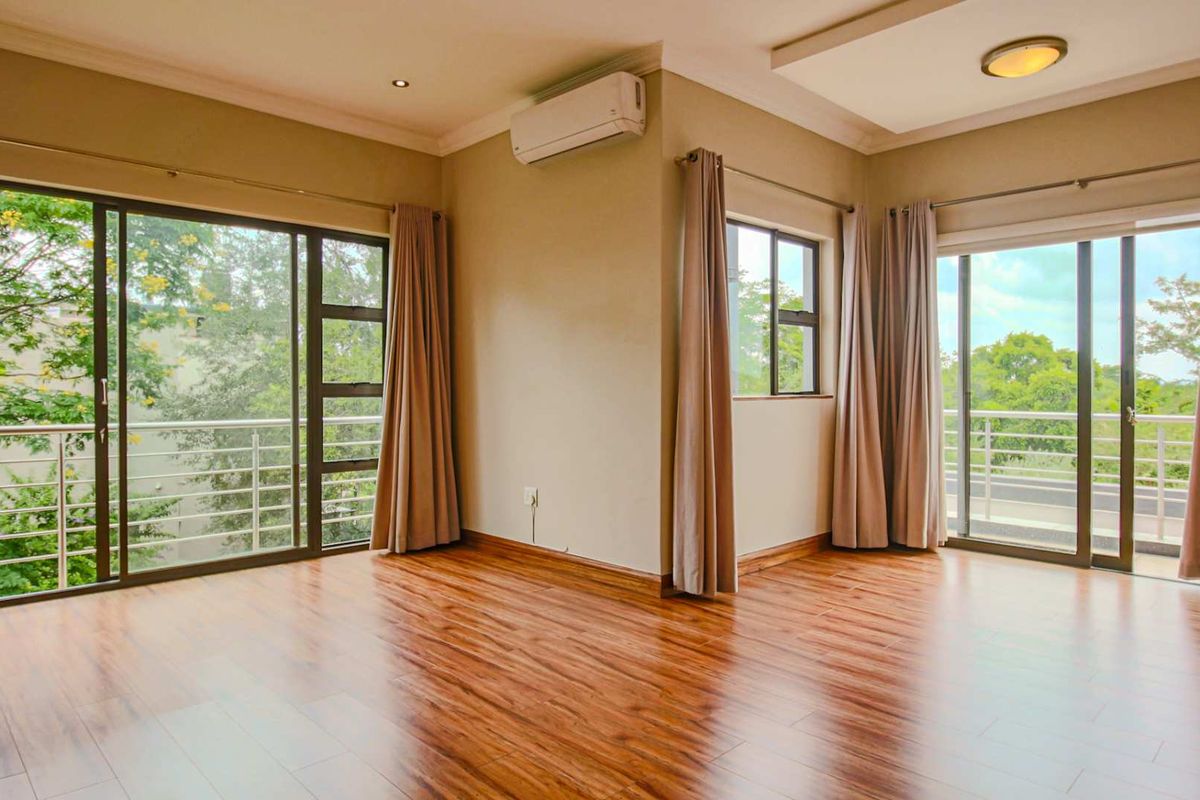 Spacious living area featuring warm wood-look flooring, recessed ceiling lighting, and dual sliding glass doors opening onto private balconies with lush green views.