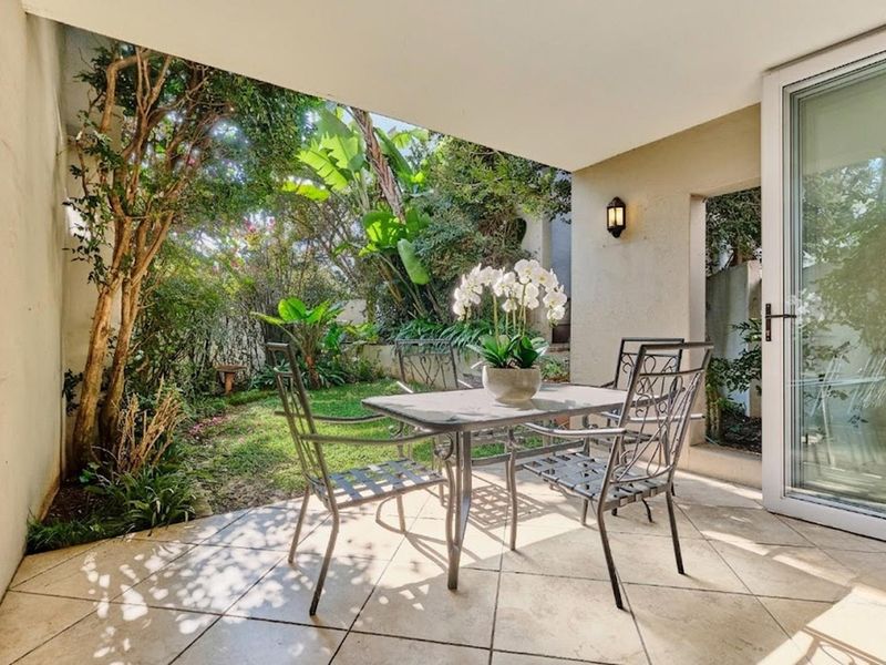 A covered patio area featuring tiled flooring and direct access to a lush, landscaped garden through a glass sliding door.