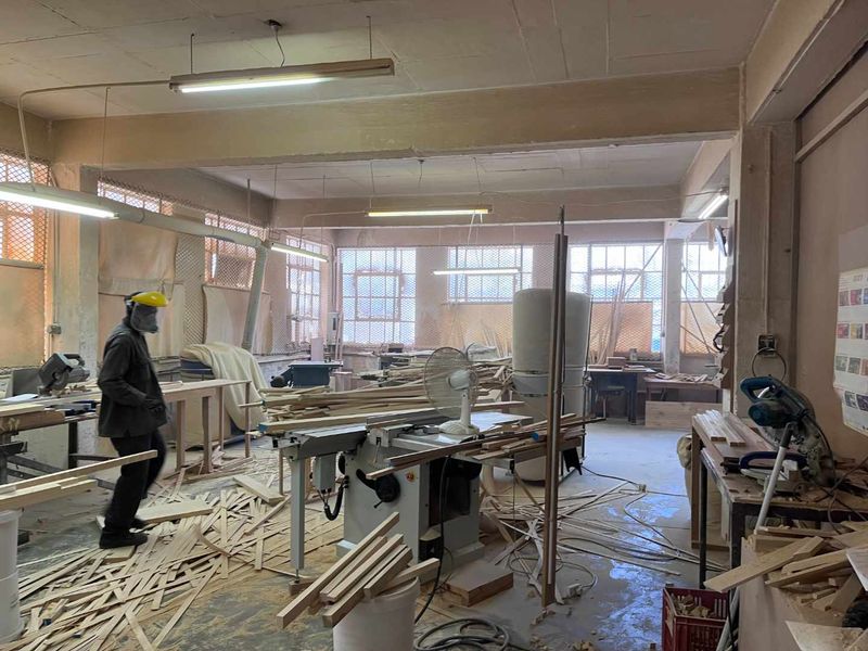 Industrial workshop space featuring exposed structural elements, large windows for natural light, and overhead fluorescent lighting, currently utilized for woodworking operations.