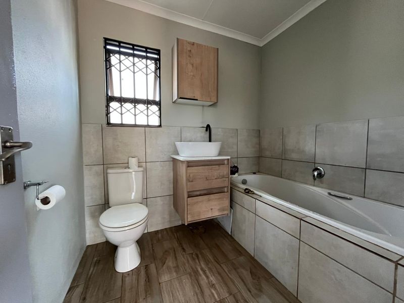 A modern bathroom featuring a floating wood-finish vanity, vessel sink, bathtub, and neutral wall and floor tiling with security bars on the window.