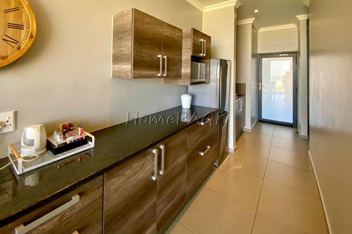 A modern galley-style kitchen featuring wood-grain cabinetry, dark stone countertops, and tiled flooring, leading to an exterior glass door.