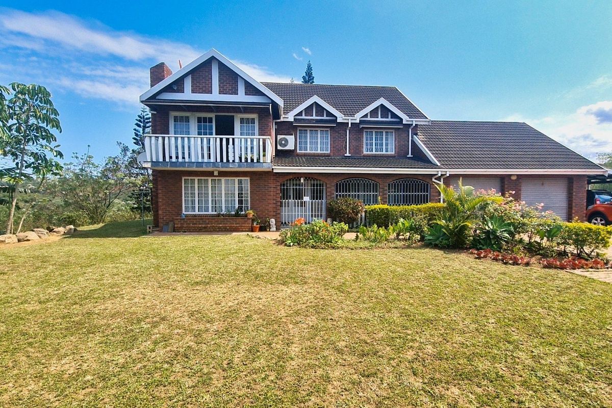 A well-maintained double-story brick house featuring a balcony, tiled roof, security gates, and an attached garage set within a spacious garden.