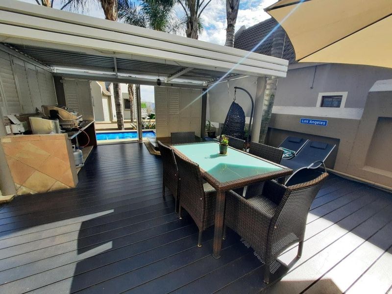 A covered outdoor patio featuring dark decking, a built-in braai area, and direct access to a swimming pool.
