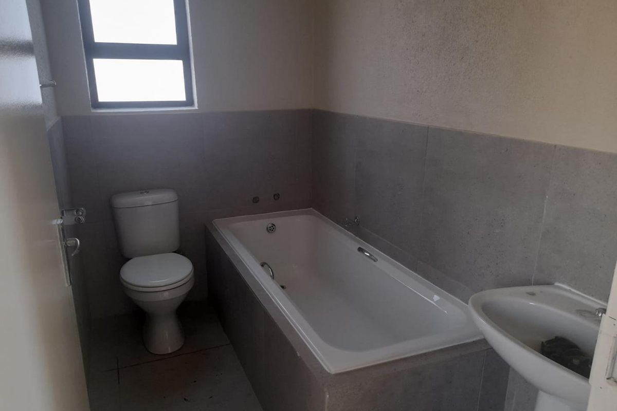 A functional bathroom featuring a built-in bathtub, pedestal basin, and toilet with neutral grey wall tiling and natural light from a window.