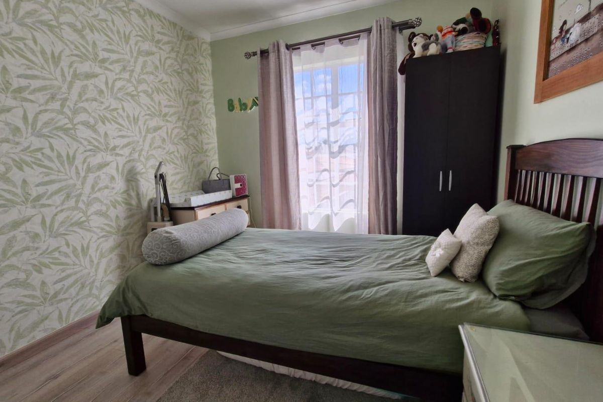 A cozy bedroom featuring wood-look laminate flooring, a decorative feature wall with leaf-patterned wallpaper, and a window with natural light.