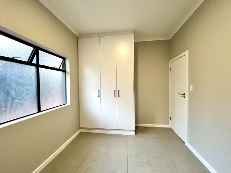A clean, empty bedroom featuring neutral tiled flooring, built-in white cupboards, and an aluminium-framed window, offering a blank canvas for personalization.