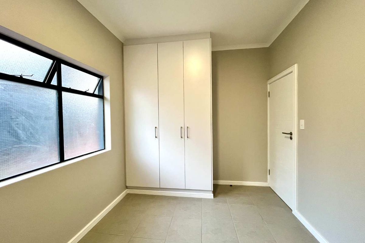 A clean, empty bedroom featuring neutral tiled flooring, built-in white cupboards, and an aluminium-framed window, offering a blank canvas for personalization.