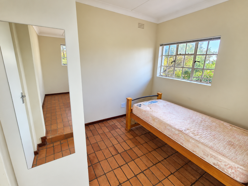 A simple bedroom featuring terracotta tile flooring, a built-in cupboard with a full-length mirror, and a window providing natural light.