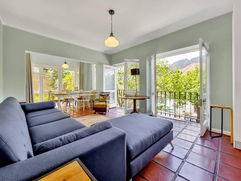 Bright open-plan living and dining area featuring terracotta flooring, pendant lighting, and multiple sets of french doors opening onto a balcony with leafy views.