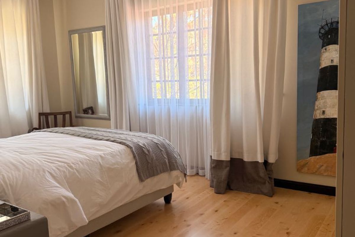 A bright and well-maintained bedroom featuring light timber flooring, a ceiling fan, and large windows dressed with elegant floor-to-ceiling curtains.