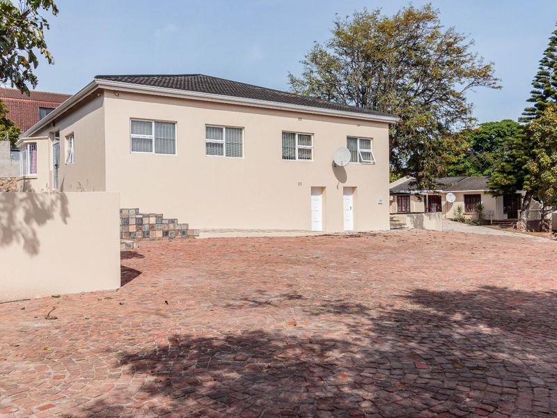 Rear view of the property featuring a large brick-paved courtyard, plastered exterior walls, and a tiled roof with established surrounding trees.