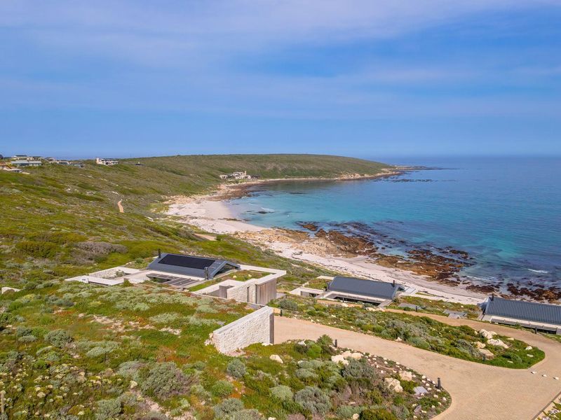 Aerial view of a modern coastal property featuring stone walls, metal roofing, solar panels, and a paved driveway overlooking the ocean.