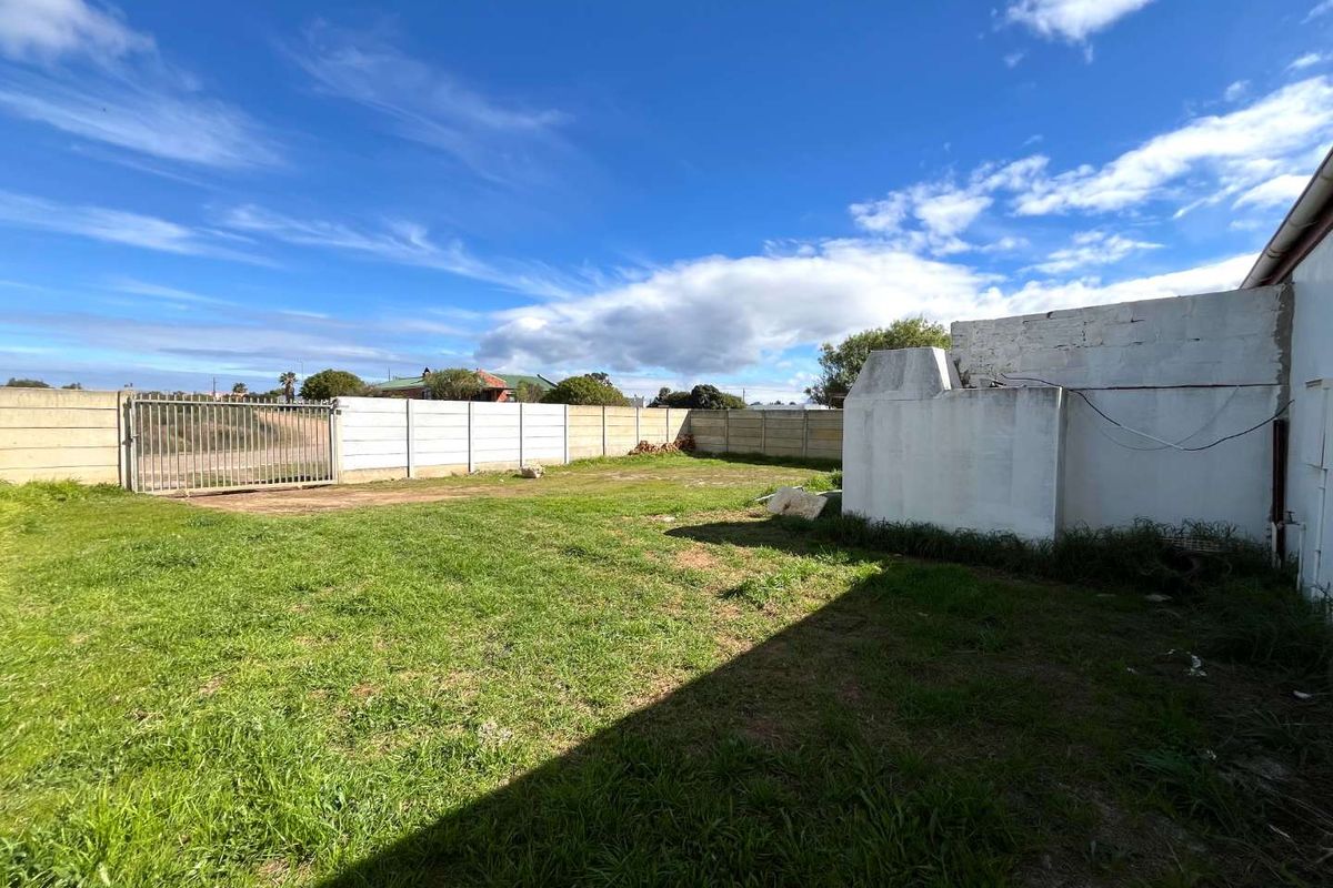 A spacious rear yard featuring a grass lawn, enclosed by a precast concrete boundary wall with a metal security gate and an existing masonry structure.