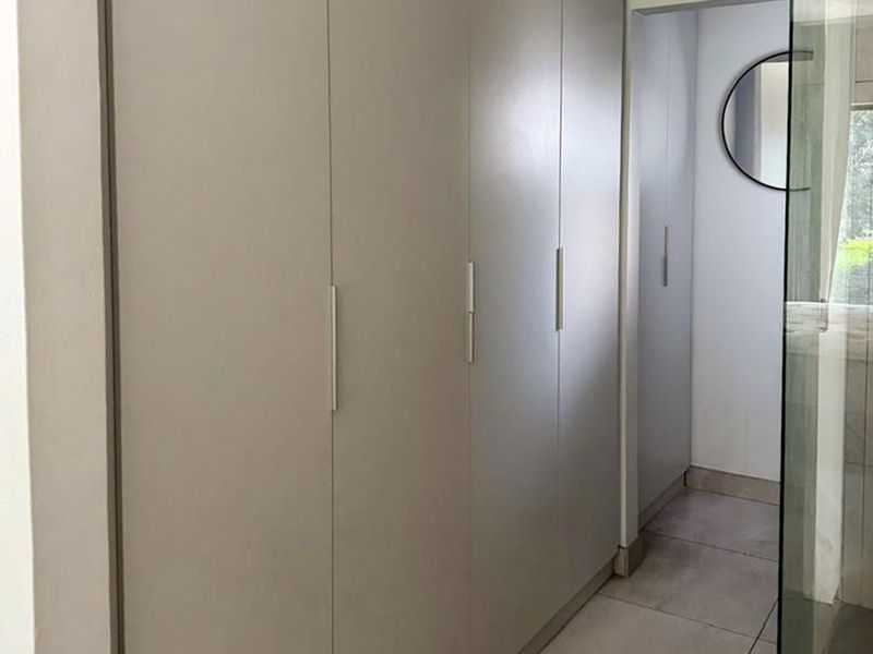 A modern bedroom area featuring floor-to-ceiling built-in cupboards, neutral tiled flooring, and an adjacent glass-enclosed shower.
